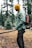 A person stands in a forested area, wearing a yellow beanie, green jacket, and dark pants. They have a colorful backpack and carry an axe attached to their belt. The background features evergreen trees and the ground is covered with light patches of snow and fallen branches.