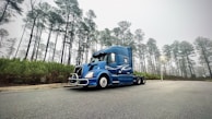A smiling truck driver standing beside his rig with a backdrop of pine trees typical of the Southeast region.