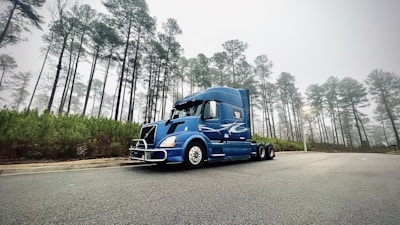 A blue semi-truck is parked on an empty road surrounded by tall pine trees. The sky is overcast, contributing to a foggy and serene atmosphere. The road appears wet, possibly from recent rain, adding a reflective surface.