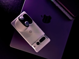 A smartphone and earbuds are placed on top of a laptop, creating a modern tech setup. The laptop has a sleek, matte finish with a subtle apple logo visible. The smartphone is aligned diagonally and the earbuds rest in their charging case.