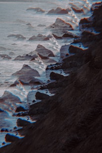 A rocky coastline with waves crashing against the shore, featuring a glitch or distortion effect that creates a surreal atmosphere. The image looks moody with its dark hues.