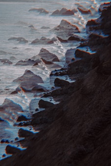 A rocky coastline with waves crashing against the shore, featuring a glitch or distortion effect that creates a surreal atmosphere. The image looks moody with its dark hues.