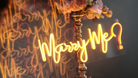 A vibrant neon sign reading 'Will U Marry Me' hanging amidst a delicate flower arch on the sand.