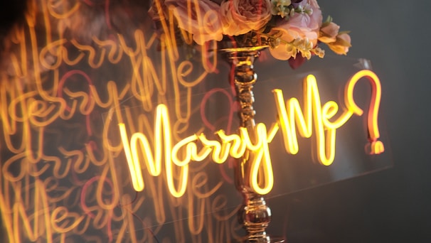 A bright neon 'Will U Marry Me' sign glowing on a beach backdrop at twilight.