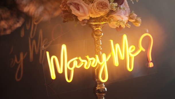 A warmly lit scene features a golden neon sign displaying the words 'Marry Me?' in elegant script. Behind the sign, a bouquet of soft pink and cream roses with small white flowers adorns a tall, shiny brass stand.