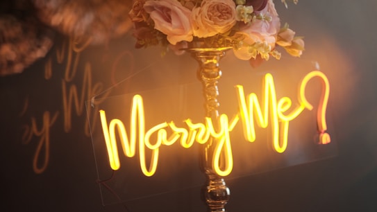 A warmly lit scene features a golden neon sign displaying the words 'Marry Me?' in elegant script. Behind the sign, a bouquet of soft pink and cream roses with small white flowers adorns a tall, shiny brass stand.