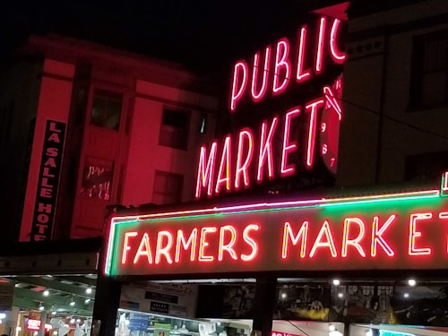 Neon signs illuminate the night, displaying 'PUBLIC MARKET' and 'FARMERS MARKET' in bright pink and green lights. Adjacent buildings are visible in the background, including a sign for 'LA SALLE HOTEL'. Below the signs, there are storefronts with various lights and details.