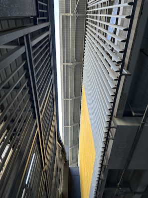 A vertical perspective of a modern industrial or architectural environment featuring metal structures. The image includes a close-up of metal beams and railings, with a large wall on the right side that has vertical slats and a yellow section.
