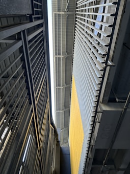 A vertical perspective of a modern industrial or architectural environment featuring metal structures. The image includes a close-up of metal beams and railings, with a large wall on the right side that has vertical slats and a yellow section.