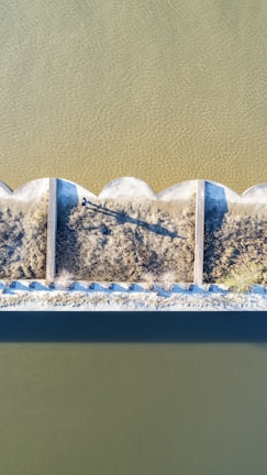 An aerial view showing a geometric structure dividing two bodies of water, with clear shadows indicating sunlight. Sparse vegetation grows along the division lines.