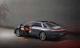 A sleek, modern car with its driver's side door open, situated in a minimalistic concrete environment. The vehicle features a futuristic design with smooth lines and an illuminated rear light strip. The interior is subtly lit, highlighting the steering wheel and dashboard.