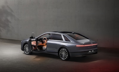 A sleek, modern car with its driver's side door open, situated in a minimalistic concrete environment. The vehicle features a futuristic design with smooth lines and an illuminated rear light strip. The interior is subtly lit, highlighting the steering wheel and dashboard.