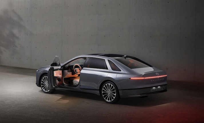 A sleek, modern car with its driver's side door open, situated in a minimalistic concrete environment. The vehicle features a futuristic design with smooth lines and an illuminated rear light strip. The interior is subtly lit, highlighting the steering wheel and dashboard.