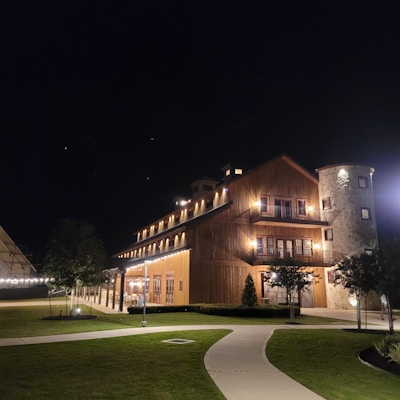 A large, rustic building is illuminated at night, featuring wooden paneling and a distinct cylindrical stone section on the right. The warm lighting accentuates the structure&rsquo;s architectural features, while a neatly manicured lawn with paved walkways leads up to it.