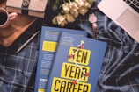 A cozy arrangement featuring a blue checkered fabric backdrop with a book titled 'Ten Year Career' prominently placed in the center. On the left, there's a beige and black handbag, next to a small red-handled mug. To the right of the book is a white wristwatch with a brown strap resting beside some pale pink roses. A silver pen lies nearby, and the corner of a laptop is visible at the top right.