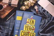 A cozy arrangement featuring a blue checkered fabric backdrop with a book titled 'Ten Year Career' prominently placed in the center. On the left, there's a beige and black handbag, next to a small red-handled mug. To the right of the book is a white wristwatch with a brown strap resting beside some pale pink roses. A silver pen lies nearby, and the corner of a laptop is visible at the top right.
