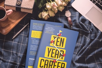 A cozy arrangement featuring a blue checkered fabric backdrop with a book titled 'Ten Year Career' prominently placed in the center. On the left, there's a beige and black handbag, next to a small red-handled mug. To the right of the book is a white wristwatch with a brown strap resting beside some pale pink roses. A silver pen lies nearby, and the corner of a laptop is visible at the top right.