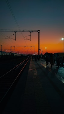 A railway station platform during sunset, with train tracks running parallel to the platform. Several people are standing and walking, casting long shadows on the ground. Overhead electrical lines and poles stretch into the distance against a vivid orange sky.