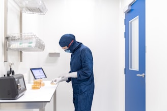 A person wearing a blue protective suit and mask is working in a laboratory setting. They are handling petri dishes on a counter with a digital display and laboratory equipment nearby. The room has clean, white walls and a blue door.