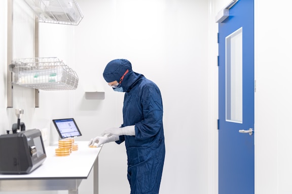 A person wearing a blue protective suit and mask is working in a laboratory setting. They are handling petri dishes on a counter with a digital display and laboratory equipment nearby. The room has clean, white walls and a blue door.