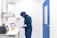A person wearing a blue protective suit and mask is working in a laboratory setting. They are handling petri dishes on a counter with a digital display and laboratory equipment nearby. The room has clean, white walls and a blue door.