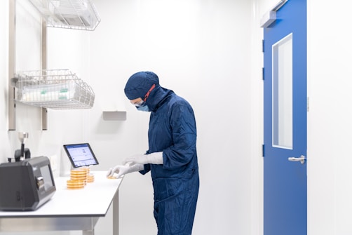 A person wearing a blue protective suit and mask is working in a laboratory setting. They are handling petri dishes on a counter with a digital display and laboratory equipment nearby. The room has clean, white walls and a blue door.