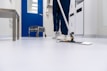 A person is mopping the floor in a clean and sterile-looking environment, possibly a hospital or laboratory. The setting includes a blue door, a cleaning cart, and stainless steel furniture.
