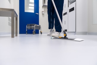 A person is mopping the floor in a clean and sterile-looking environment, possibly a hospital or laboratory. The setting includes a blue door, a cleaning cart, and stainless steel furniture.