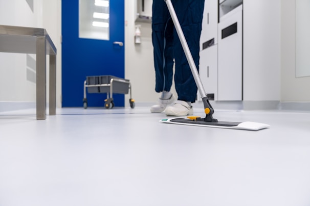 A person is mopping the floor in a clean and sterile-looking environment, possibly a hospital or laboratory. The setting includes a blue door, a cleaning cart, and stainless steel furniture.