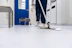 A person is mopping the floor in a clean and sterile-looking environment, possibly a hospital or laboratory. The setting includes a blue door, a cleaning cart, and stainless steel furniture.