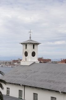 A clock tower with two black clock faces is set atop a white building with a gray shingled roof. The sky is overcast, creating a muted atmosphere. In the background, there are several buildings and a marina filled with sailboats.