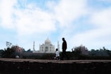 A silhouetted man and child walk on a path in the foreground, with the iconic white marble structure of the Taj Mahal prominently visible in the background. The sky is partly cloudy, creating a serene and tranquil atmosphere.