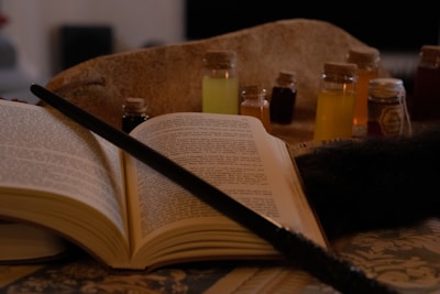 An open book rests on an intricately patterned surface, with a dark wand placed diagonally across its pages. Several small glass bottles filled with colorful liquids are arranged in the background, suggesting a magical or alchemical theme. A soft, fur-like texture partially occupies the right side of the image.