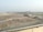 The image depicts a construction site with piles of rocks and an excavator situated near a water body. In the background, several multi-story buildings are visible, indicating an urban area. The landscape is mostly barren, with sparse greenery and a clear sky.
