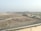 The image depicts a construction site with piles of rocks and an excavator situated near a water body. In the background, several multi-story buildings are visible, indicating an urban area. The landscape is mostly barren, with sparse greenery and a clear sky.