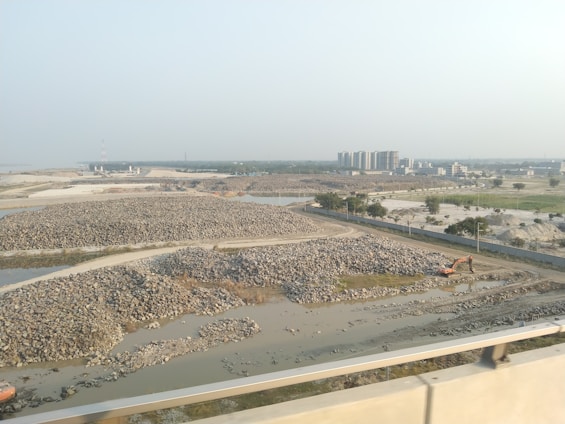 The image depicts a construction site with piles of rocks and an excavator situated near a water body. In the background, several multi-story buildings are visible, indicating an urban area. The landscape is mostly barren, with sparse greenery and a clear sky.