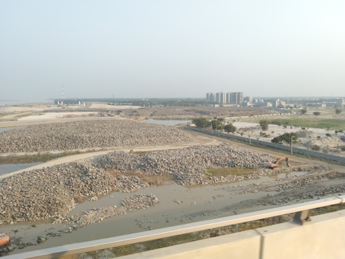 The image depicts a construction site with piles of rocks and an excavator situated near a water body. In the background, several multi-story buildings are visible, indicating an urban area. The landscape is mostly barren, with sparse greenery and a clear sky.