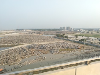 The image depicts a construction site with piles of rocks and an excavator situated near a water body. In the background, several multi-story buildings are visible, indicating an urban area. The landscape is mostly barren, with sparse greenery and a clear sky.