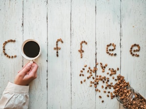 Coffee beans are meticulously arranged on a rustic wooden surface to spell out the word 'coffee.' A hand is holding a cup filled with dark coffee, forming the letter 'o.' A glass jar lies on its side, spilling coffee beans across the table.