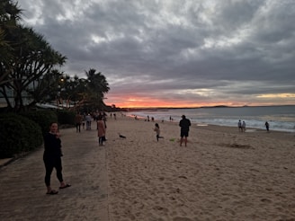 A sandy beach with several people walking and relaxing. The sky is covered with dramatic clouds, and a vibrant sunset casts hues of orange and pink. Palm trees line the walkway along the left, where additional people are strolling. The ocean waves gently lap against the shore, creating a serene atmosphere.