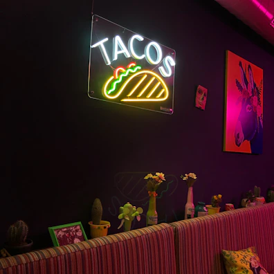 A dimly lit room features a neon sign with the word 'TACOS' and a taco graphic on a dark wall. Below, a striped bench has decorative cushions. Various potted cacti and flowers are arranged on a narrow ledge, with colorful ornaments including an artistic painting of a donkey with a bright backdrop. The atmosphere combines modern neon lighting with quirky, eclectic decor.