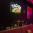 A dimly lit room features a neon sign with the word 'TACOS' and a taco graphic on a dark wall. Below, a striped bench has decorative cushions. Various potted cacti and flowers are arranged on a narrow ledge, with colorful ornaments including an artistic painting of a donkey with a bright backdrop. The atmosphere combines modern neon lighting with quirky, eclectic decor.