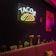 A dimly lit room features a neon sign with the word 'TACOS' and a taco graphic on a dark wall. Below, a striped bench has decorative cushions. Various potted cacti and flowers are arranged on a narrow ledge, with colorful ornaments including an artistic painting of a donkey with a bright backdrop. The atmosphere combines modern neon lighting with quirky, eclectic decor.