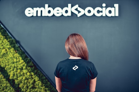 A person with long hair wearing a black t-shirt stands facing a dark wall. The shirt features a small white logo on the back. Above, white text displays the word 'embedsocial'. The left side of the image shows a section of green plant wall art.