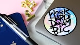 A close-up of a workspace featuring a silver laptop with a colorful sticker reading 'Work Hard Dream Big', a blue tablet with an Apple logo, a black and clear pen, pink notepad, and golden binder clips.