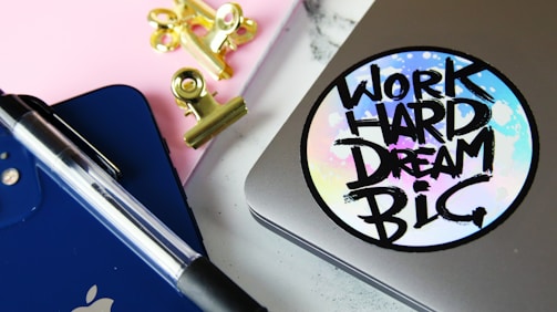 A close-up of a workspace featuring a silver laptop with a colorful sticker reading 'Work Hard Dream Big', a blue tablet with an Apple logo, a black and clear pen, pink notepad, and golden binder clips.