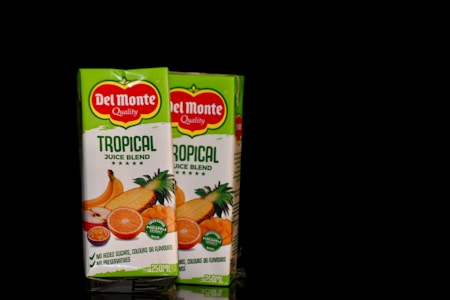 Two boxes of Del Monte Tropical Juice Blend are displayed against a black background. The packaging is predominantly green with images of tropical fruits like oranges, pineapples, and bananas. The label emphasizes quality with notes about no added sugars or preservatives.