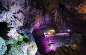 A dimly lit cave with rugged rock formations and vibrant lighting effects. The lighting creates an array of colors including purple, green, and yellow, highlighting the textures and contours of the cave walls. Stairs descend into the cave, enhancing the mysterious and adventurous atmosphere. The cave’s ceiling is high and shadowy, adding depth and intrigue.