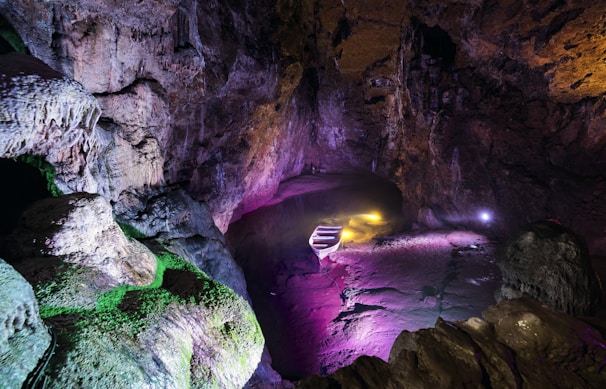 A dimly lit cave with rugged rock formations and vibrant lighting effects. The lighting creates an array of colors including purple, green, and yellow, highlighting the textures and contours of the cave walls. Stairs descend into the cave, enhancing the mysterious and adventurous atmosphere. The cave’s ceiling is high and shadowy, adding depth and intrigue.