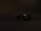A dark, sleek sports car is positioned against a plain black background, highlighting its aerodynamic design and smooth curves. The lighting is minimal, casting subtle shadows and emphasizing the car's polished surface.